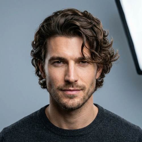 A close-up studio portrait of a 29-year-old man with stylish, voluminous wavy hair and a stubble beard.